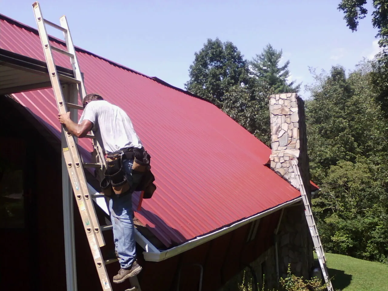 Roofer on a ladder beside a red metal roof and stone chimney — Soffit & Fascia Repair by Roofing Brothers in Ouray